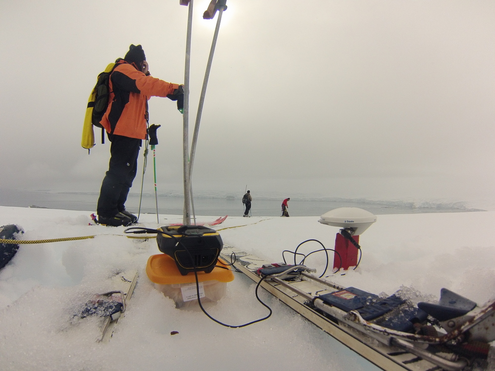 Антарктика, GPS приемник в режим на статично измерване върху дървен репер в ледника Перуника, в дъното - залива Емона. На снимката – Цецо (преден план), Божо и Васил
